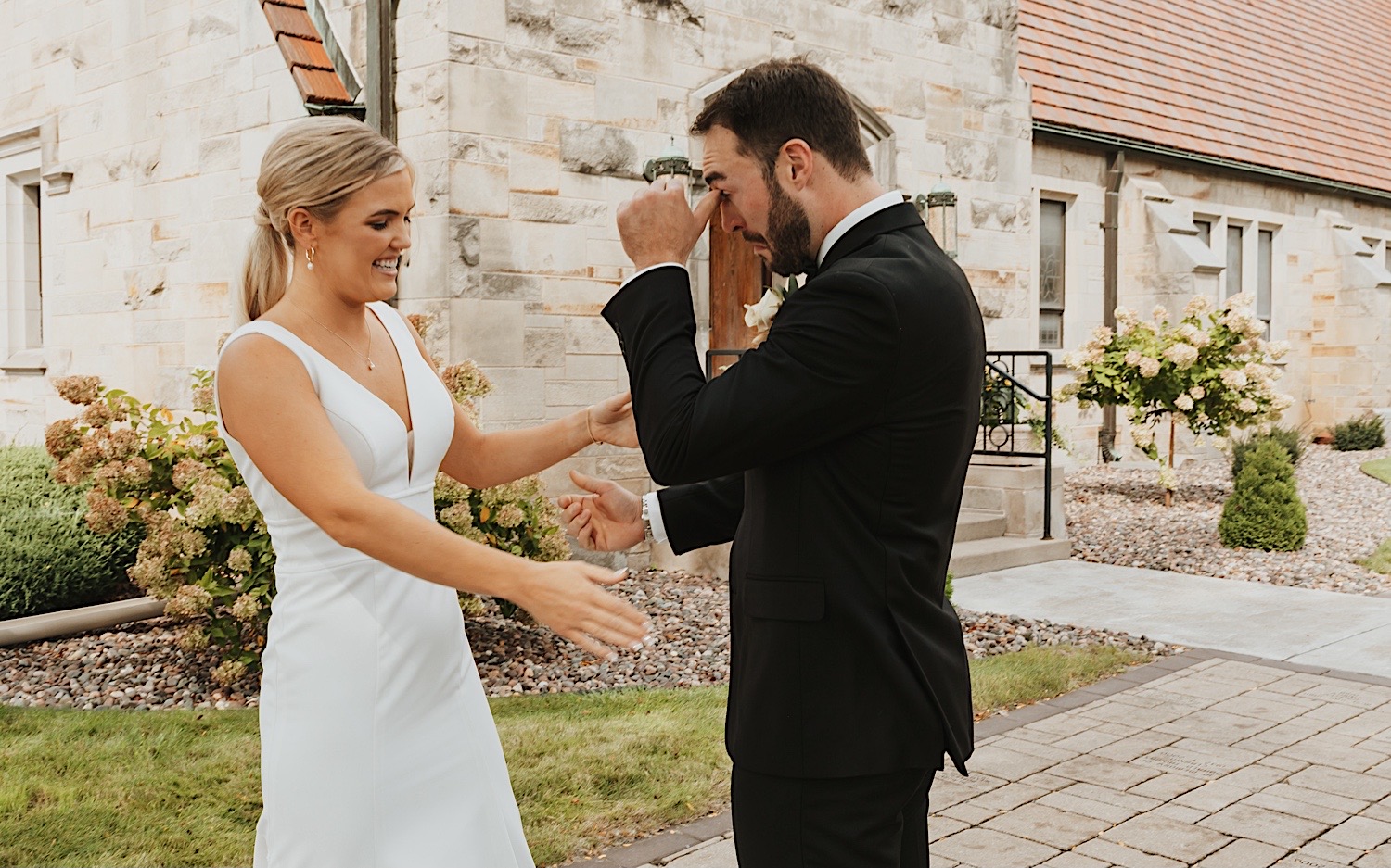 A groom wipes tears from his face as his bride smiles at him during their first look