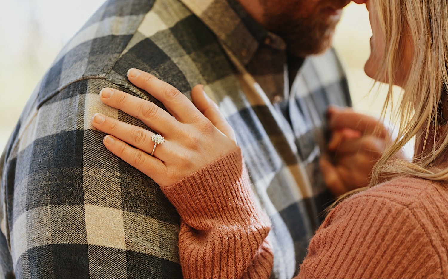 Close up photo of a woman's hand with an engagement ring resting on a man's shoulder as they kiss off camera