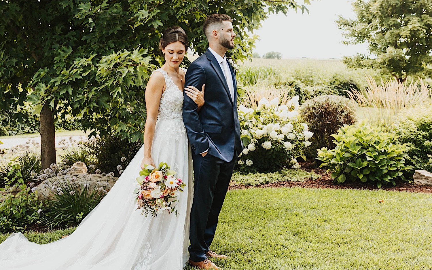 A bride stands behind the groom and locks arms with him while they stand outdoors at their wedding venue Legacy Hill Farm