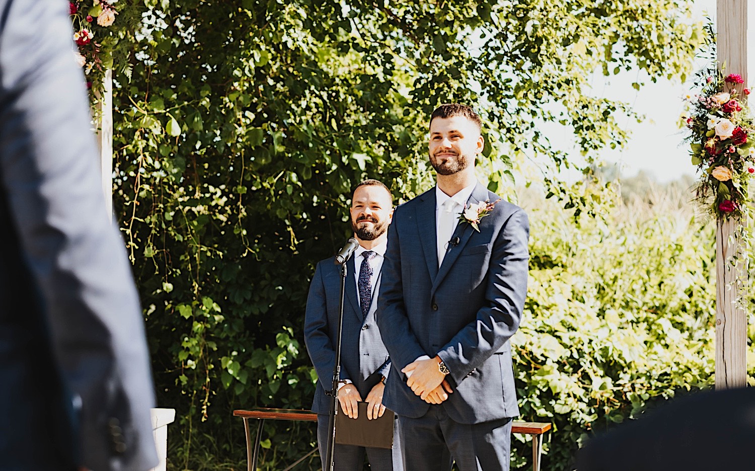 A groom stands and smiles with hands clasped while at the outdoor ceremony space of the wedding venue Legacy Hill Farm