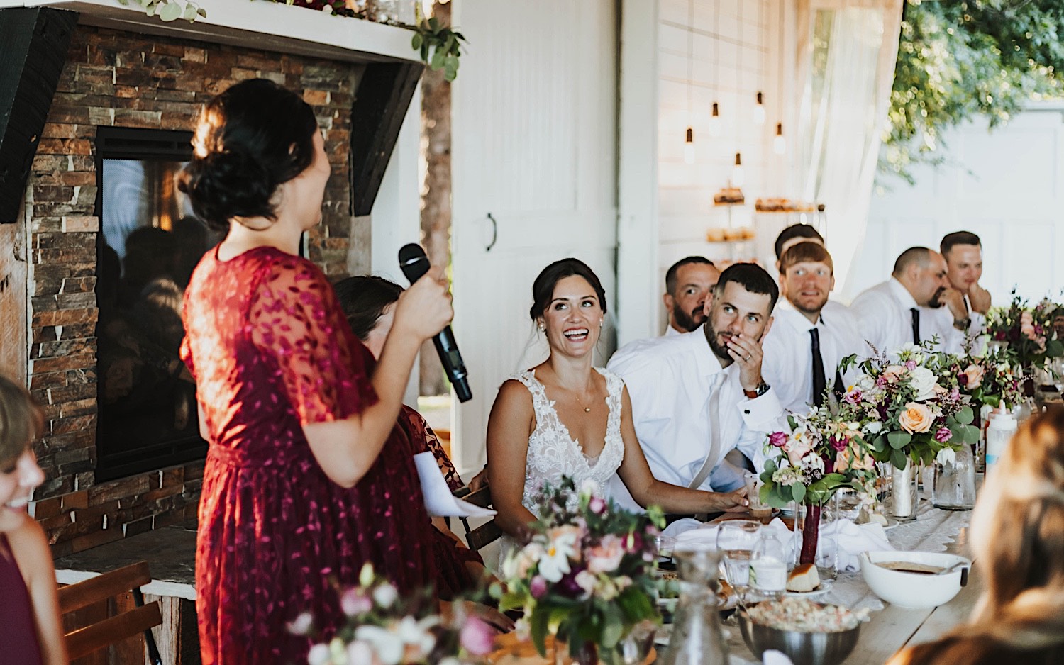 A bride smiles while sitting and listening to a bridesmaid give a speech during her wedding reception at Legacy Hill Farm